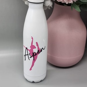 May include: White stainless steel water bottle with a pink silhouette of a dancer and the name "Aspen" in black lettering.