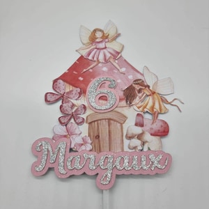 May include: A pink and white fairy cake topper with a mushroom house, two fairies, and the name "Margaux" and the number "6" in silver glitter.