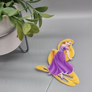 May include: A paper cutout of Rapunzel from the Disney film Tangled. She is wearing a purple dress and has long blonde hair.