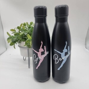 May include: Two black water bottles with a dancer silhouette and personalized names. The bottle on the left says "Grace" in pink lettering and the bottle on the right says "Bonnie" in blue lettering.