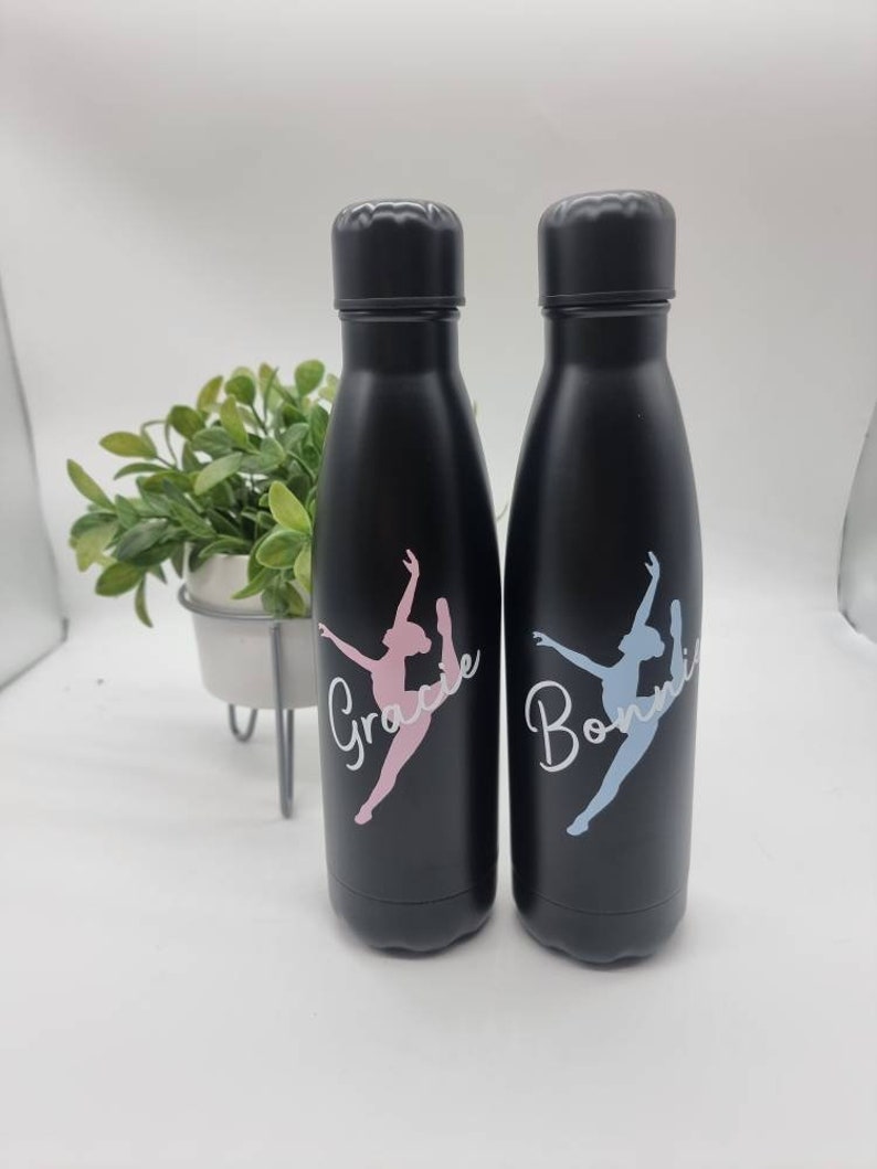 May include: Two black water bottles with a dancer silhouette and personalized names. The bottle on the left says "Grace" in pink lettering and the bottle on the right says "Bonnie" in blue lettering.