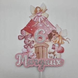 May include: A pink and red fairy house cake topper with a silver glitter number 6 and the name Margaux in silver glitter. The topper features two fairies with wings and a mushroom.