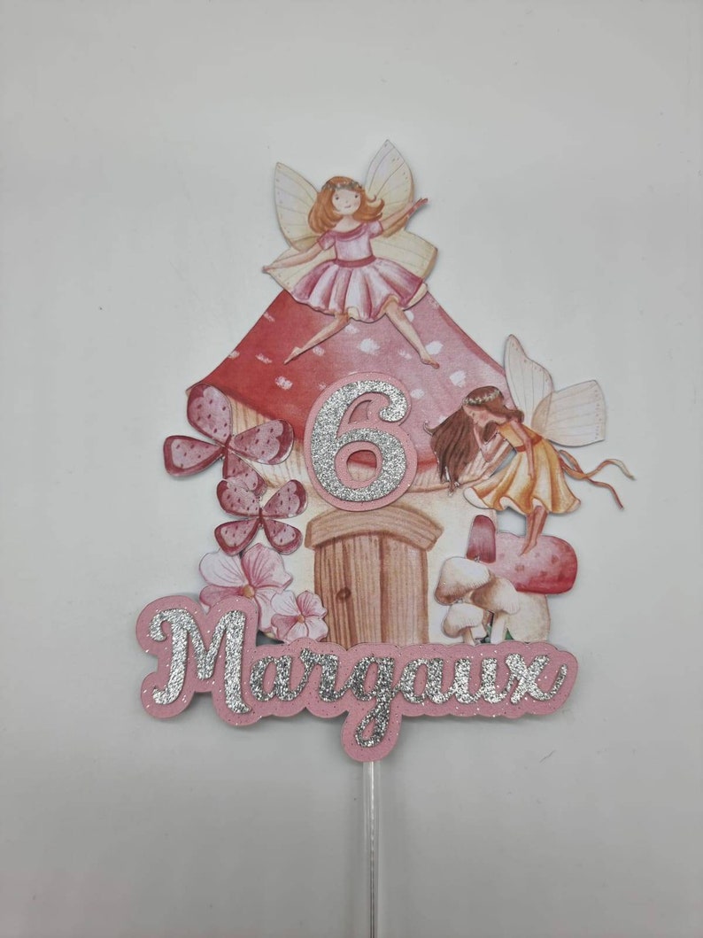 May include: A pink and red fairy house cake topper with a silver glitter number 6 and the name Margaux in silver glitter. The topper features two fairies with wings and a mushroom.