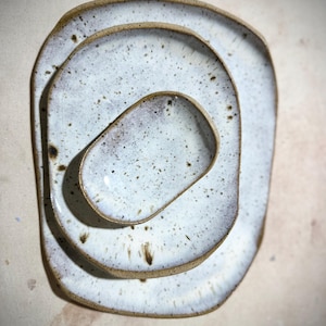 May include: A stack of three ceramic plates with a speckled white glaze and a brown rim. The plates are oval-shaped with rounded corners, nested inside each other. The smallest plate is a shallow dish.