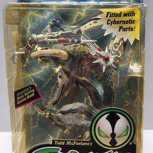 Op de afbeelding: Een McFarlane Toys Violator II actiefiguur in de originele blisterverpakking. Het wezen heeft een getextureerde bruine huid, grote hoorns en zilveren cybernetische onderdelen. De verpakking bevat het Spawn-logo, de tekst Fitted with Cybernetic Parts! en America's Best Selling Comic Book.