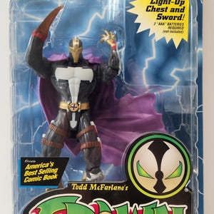 Puede incluir: Figura de acción Redeemer de la edición Deluxe de la serie de cómics Spawn. La figura está en un blíster con el texto "Light-Up Chest and Sword!" y "Ultra-Action Figures". La figura es negra, dorada y morada.