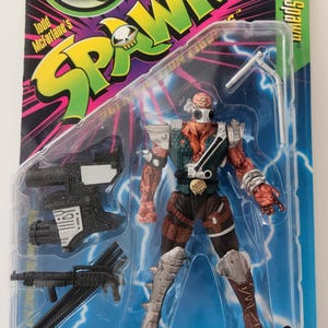 Op de afbeelding: Een Spawn actiefiguur in een plastic verpakking. De figuur is een gespierd personage met een rood en groen kleurenschema, gekleed in een harnas en met wapens. De verpakking toont het Spawn-logo en de tekst "Giant Weapon Fires Rat Missile!"