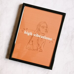 May include: A framed print with a peach background featuring a line drawing of a woman's face and the text "high vibrations".