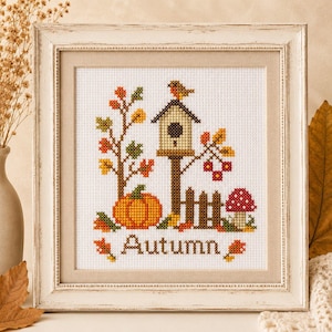 Könnte beinhalten: Ein gerahmtes Kreuzstich-Stickbild mit einer Herbstszene, bestehend aus einem Vogelhaus, einem Vogel, einem Kürbis, einem Pilz und einem Holzzaun. Das Motiv zeigt buntes Herbstlaub und das Wort Autumn in braunem Garn auf weißem Stoff.