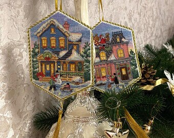May include: Two hand-stitched Christmas ornaments featuring winter scenes. The ornaments are shaped like hexagons and have a gold tassel. The first ornament depicts a blue house with snow on the roof and a family outside. The second ornament depicts a pink house with Santa Claus on the roof.