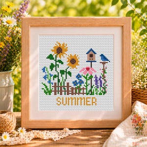 May include: A framed cross-stitch featuring a colorful summer garden scene. The design includes sunflowers, a birdhouse, a bluebird, and the word "SUMMER" in gold lettering. The frame is made of light brown wood.