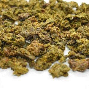 May include: Close-up of a pile of dried, green and brown propolis granules. The granules are irregularly shaped and have a rough texture.