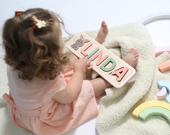 May include: A wooden puzzle with the name "LINDA" spelled out in colorful letters. The puzzle is shaped like a rectangle and has a small bunny rabbit cutout in the top left corner. A young child is holding the puzzle in their hands.