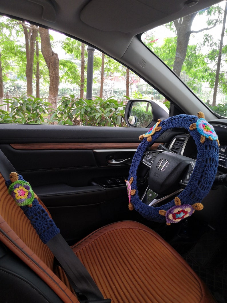 May include: A blue crocheted steering wheel cover with colorful turtle designs. The cover is installed on a car's steering wheel. A matching crocheted turtle design is on the seatbelt.