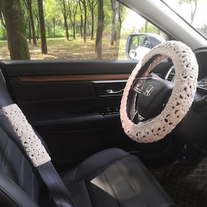 May include: A white crocheted steering wheel cover and a white crocheted seatbelt cover in a car interior. The car has black leather seats and a wooden steering wheel.