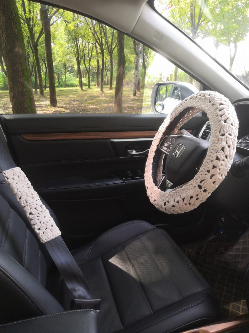 May include: A white crocheted steering wheel cover and a white crocheted seatbelt cover in a car interior. The car has black leather seats and a wooden steering wheel.