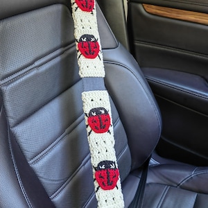 May include: A beige crochet seat belt cover featuring four red and black ladybug motifs. The rectangular cover is wrapped around a gray car seat belt, displaying a repeating pattern of the insects with black heads and red wings.