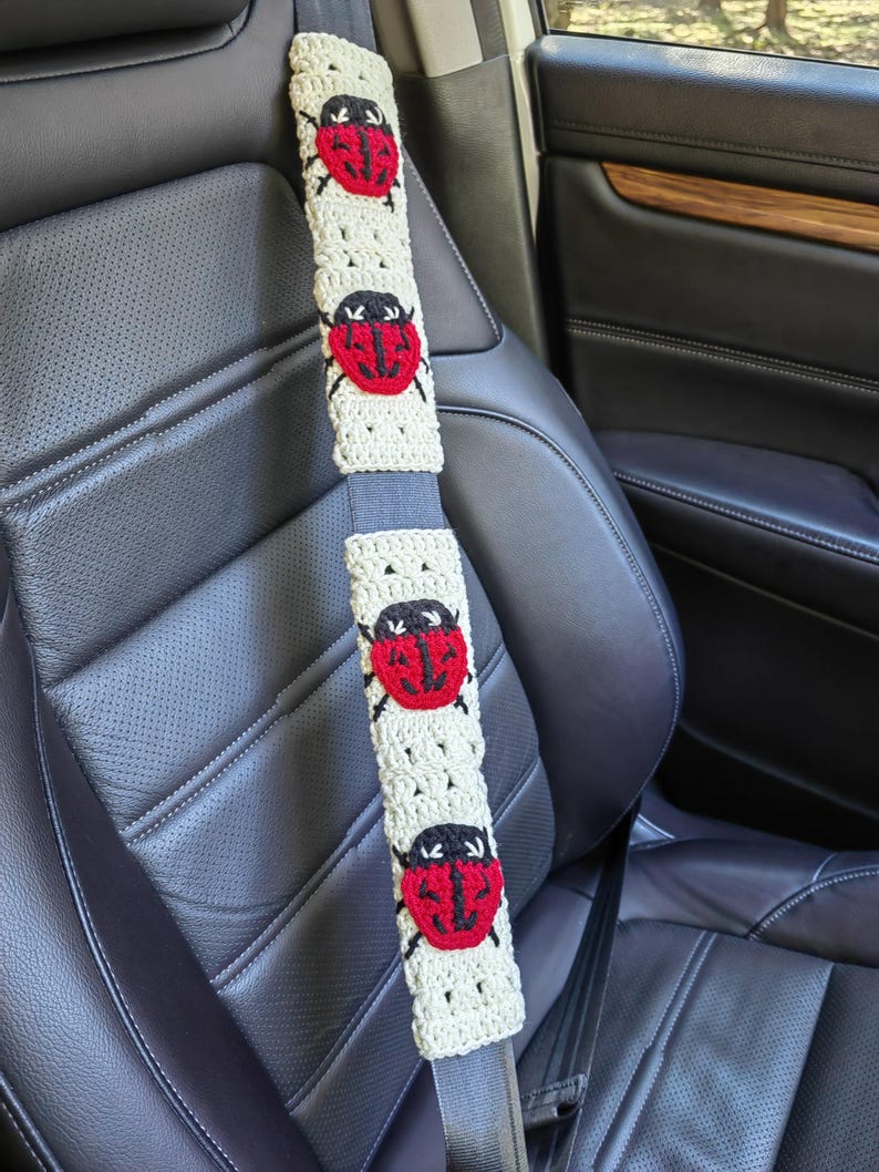 May include: A beige crochet seat belt cover featuring four red and black ladybug motifs. The rectangular cover is wrapped around a gray car seat belt, displaying a repeating pattern of the insects with black heads and red wings.