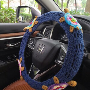 May include: A blue crocheted steering wheel cover with colorful crocheted turtles attached. The cover is on a car steering wheel.