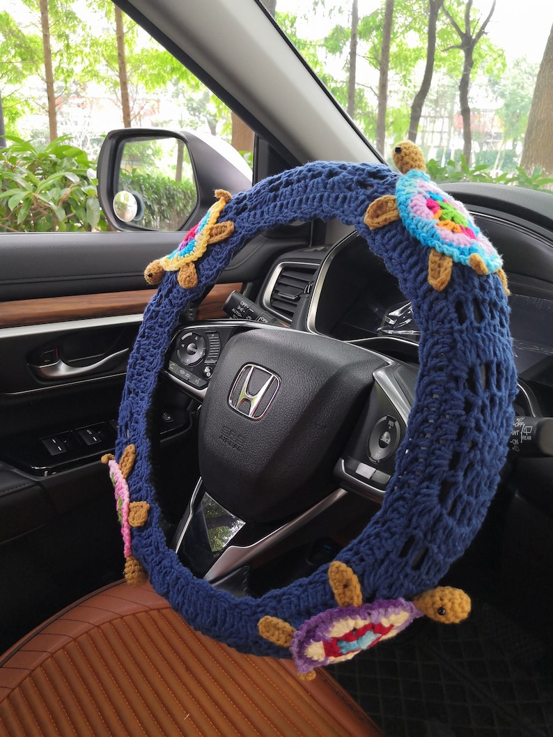 May include: A blue crocheted steering wheel cover with colorful crocheted turtles attached. The cover is on a car steering wheel.