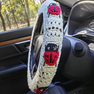 May include: A cream-colored crocheted steering wheel cover featuring a repeating pattern of red and black ladybug motifs. The cover is constructed with a textured yarn stitch and fits snugly around the circular rim of the steering wheel.