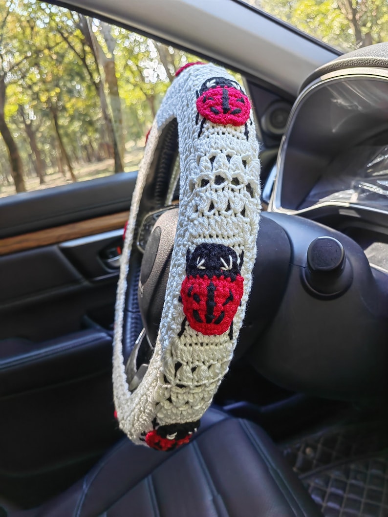 May include: A cream-colored crocheted steering wheel cover featuring a repeating pattern of red and black ladybug motifs. The cover is constructed with a textured yarn stitch and fits snugly around the circular rim of the steering wheel.