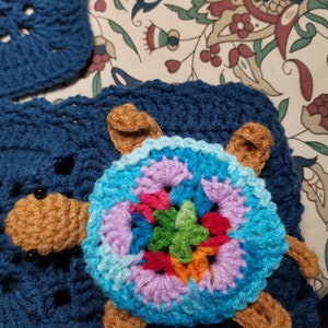 May include: A crocheted turtle with a colorful shell and brown legs. The turtle is sitting on a blue crocheted blanket.