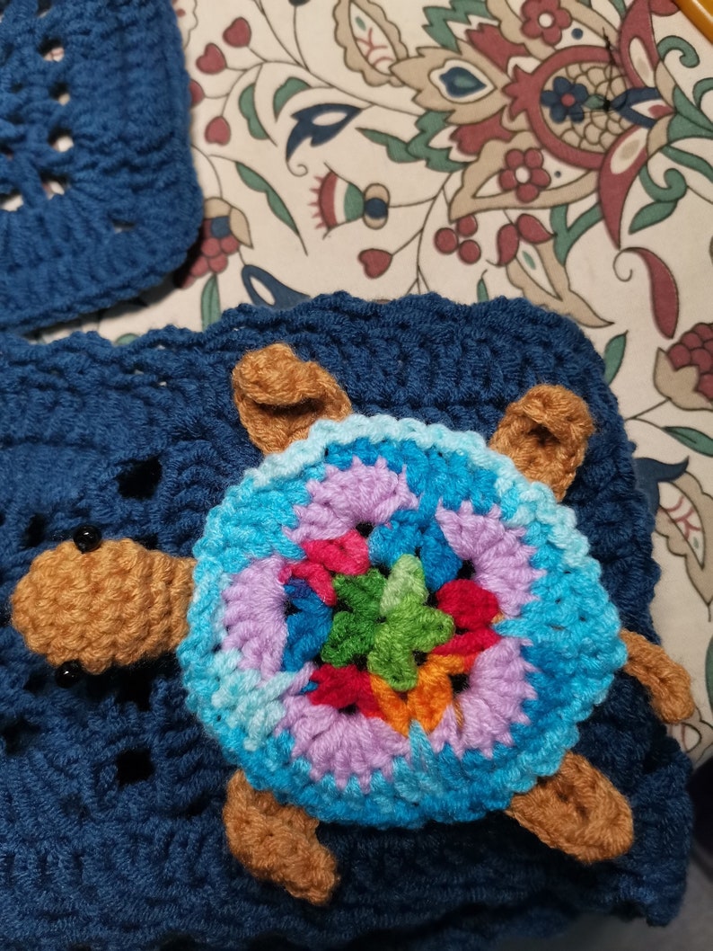 May include: A crocheted turtle with a colorful shell and brown legs. The turtle is sitting on a blue crocheted blanket.