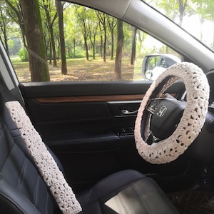 May include: White crocheted steering wheel cover and seatbelt cover in a car interior. The car is parked in a forest with green trees visible through the window.