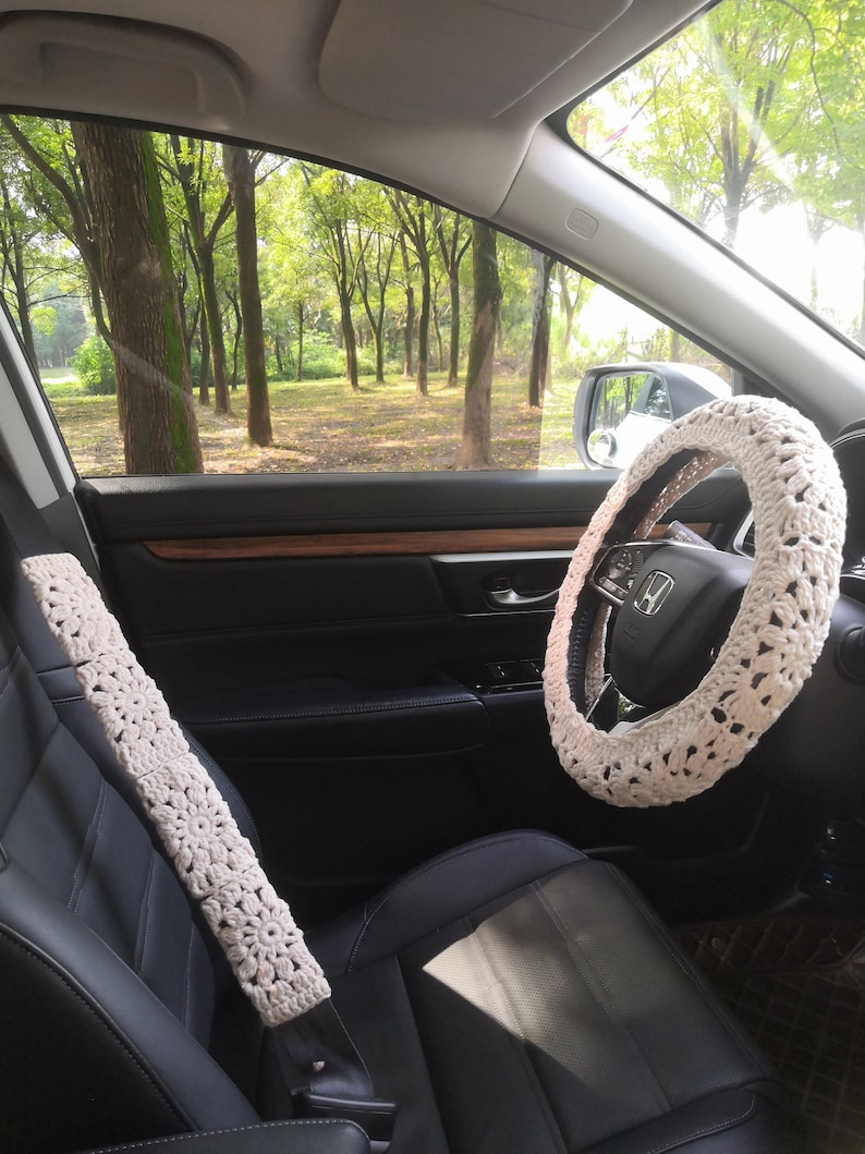 May include: White crocheted steering wheel cover and seatbelt cover in a car interior. The car is parked in a forest with green trees visible through the window.
