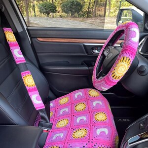 May include: Crocheted car accessories in pink, yellow, and white. The set includes a seat cover, steering wheel cover, and seatbelt covers, all featuring a sunflower design. The interior of the car is black with wood trim, and the background shows trees.
