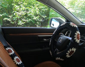May include: Interior car accessories, including a black steering wheel cover and seat belt cover, both adorned with a crochet daisy pattern in white, brown, and black. The car seat is brown.