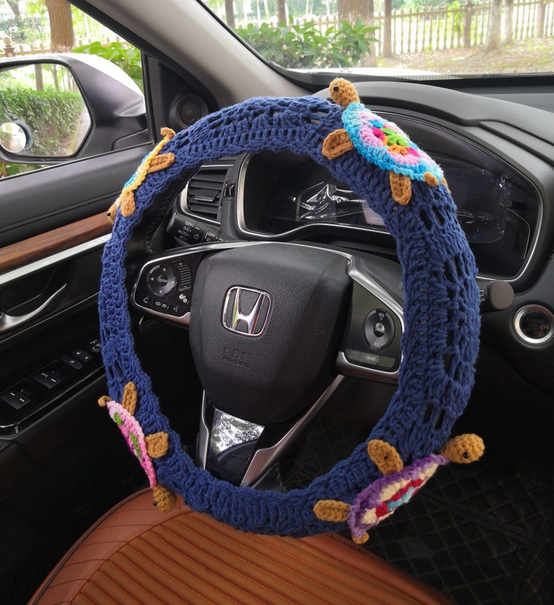 May include: A blue crocheted steering wheel cover with colorful crocheted turtles attached. The cover is on a car steering wheel.