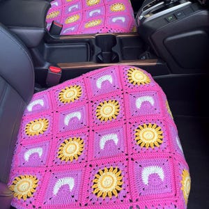 May include: Handmade crochet car seat covers in a vibrant pink, featuring a repeating pattern of yellow sunflowers and white crescent shapes. The covers are placed on the front seats of a car, adding a touch of handmade charm to the interior.