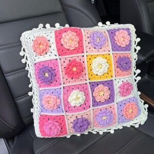 Puede incluir: Una almohada cuadrada de crochet con un borde blanco y un patrón floral en tonos de rosa, morado y amarillo. La almohada está sentada en un asiento de coche de cuero negro.