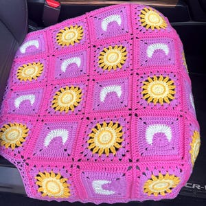 May include: A handmade crochet car seat cover in a vibrant pink. The design features square motifs with yellow sunflowers, white crescent shapes, and purple accents. The cover is made of soft yarn and adds a touch of color to the car interior.
