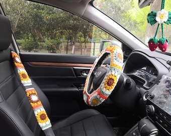 May include: A crocheted steering wheel cover with a floral pattern in shades of yellow, orange, white, and green. The cover is installed on a black leather steering wheel in a car interior.