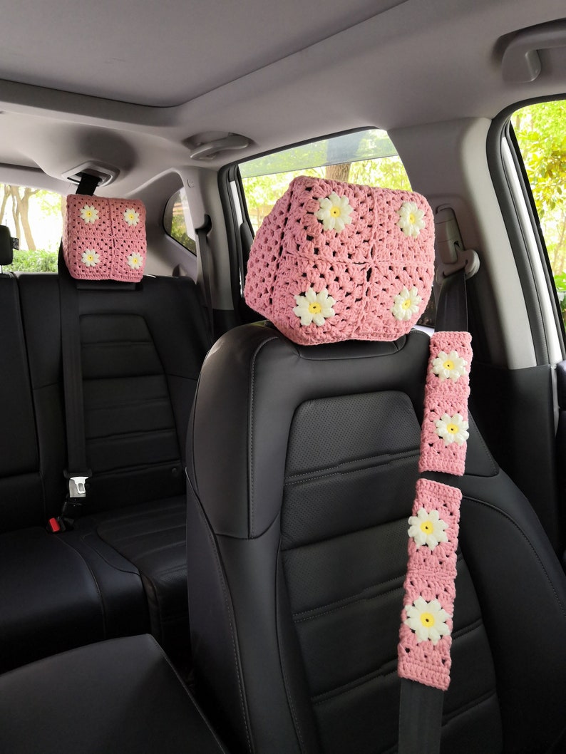 May include: Pink crocheted car seat headrest covers with white daisy accents. The headrest covers are shown on the front seats of a car. The seatbelt is also covered with a pink crocheted cover with white daisy accents.