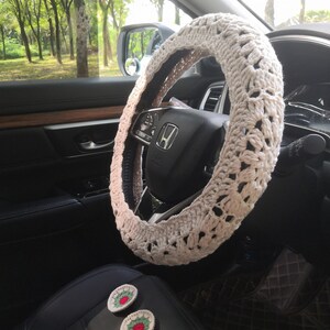 May include: A white crocheted steering wheel cover with a delicate pattern. The cover is installed on a black leather steering wheel in a car interior. Two small crocheted flowers with red and green accents are visible on the passenger seat.