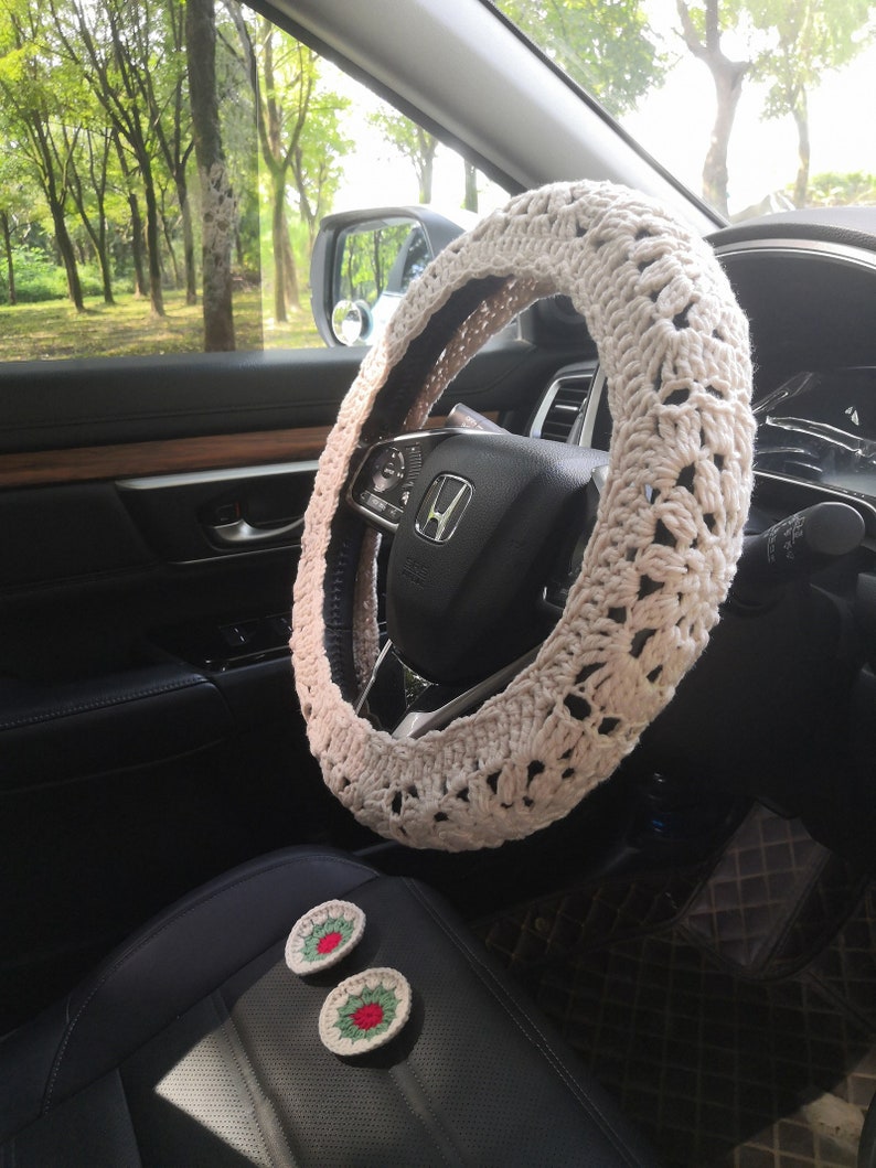May include: A white crocheted steering wheel cover with a delicate pattern. The cover is installed on a black leather steering wheel in a car interior. Two small crocheted flowers with red and green accents are visible on the passenger seat.