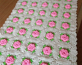 Könnte beinhalten: Eine weiße Häkeldecke mit einem sich wiederholenden Muster aus rosa Rosen und grünen Blättern.