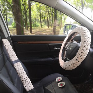 May include: A car interior with black leather seats, a white crocheted steering wheel cover, and a white crocheted seatbelt cover. There are two small red and green embroidered flower decorations on the passenger seat.