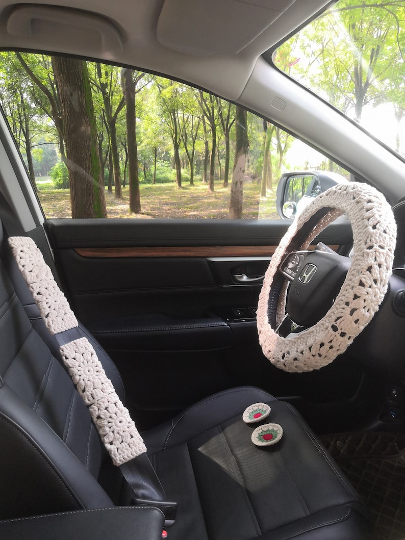 May include: A car interior with black leather seats, a white crocheted steering wheel cover, and a white crocheted seatbelt cover. There are two small red and green embroidered flower decorations on the passenger seat.