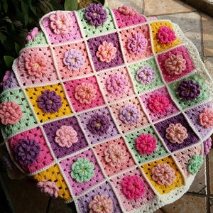 May include: A crocheted baby blanket with a floral design in shades of pink, purple, green, and yellow. The blanket is made up of individual squares with a flower in the centre of each square.