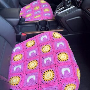 May include: Pink crochet car seat covers with a sunflower design. The covers feature a pattern of yellow sunflowers, white arches, and pink squares. The covers are placed on the front seats of a car.