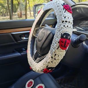 May include: A cream-colored crocheted steering wheel cover featuring repeating red and black ladybug motifs. The cover is fitted onto a black steering wheel, and two small, round, red and cream crocheted coasters are placed on the seat below.