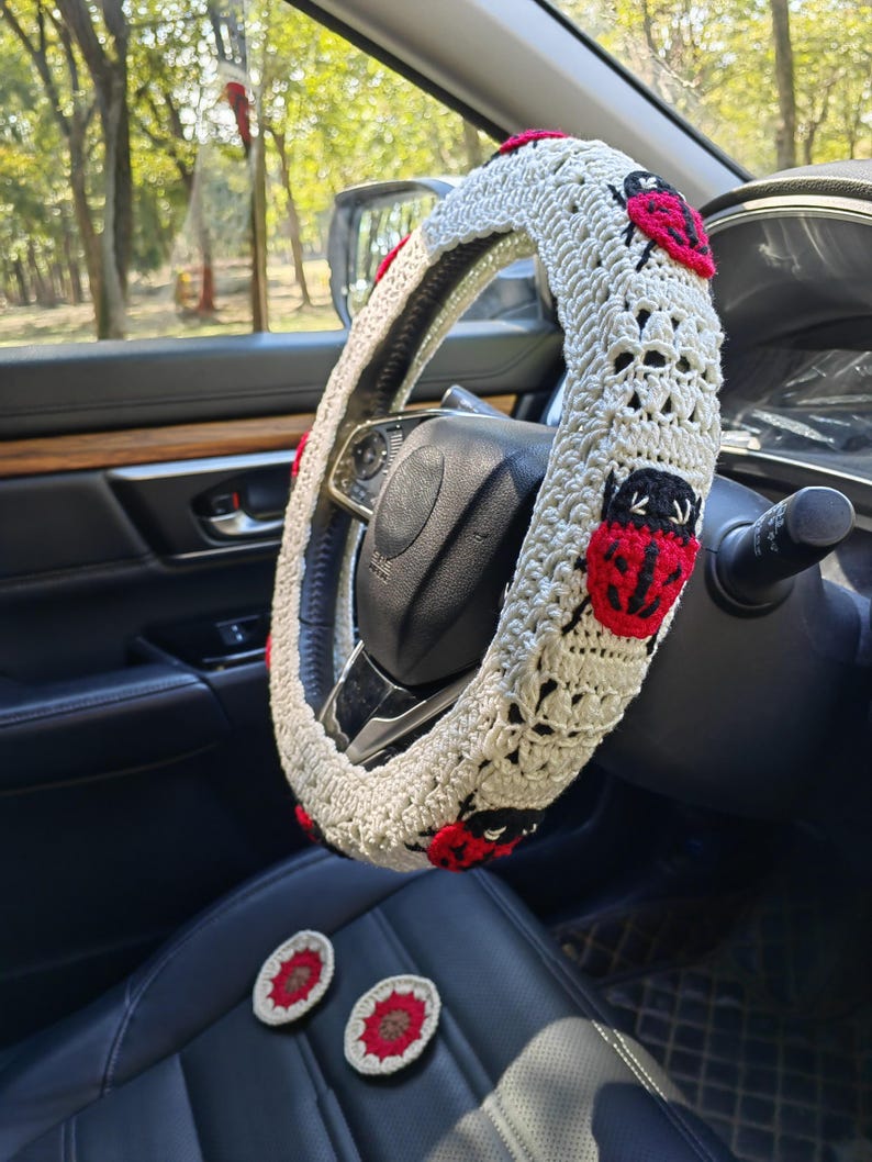 May include: A cream-colored crocheted steering wheel cover featuring repeating red and black ladybug motifs. The cover is fitted onto a black steering wheel, and two small, round, red and cream crocheted coasters are placed on the seat below.