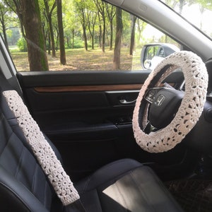 May include: A white crocheted steering wheel cover and a white crocheted seatbelt cover in a car interior. The car is parked in a wooded area.