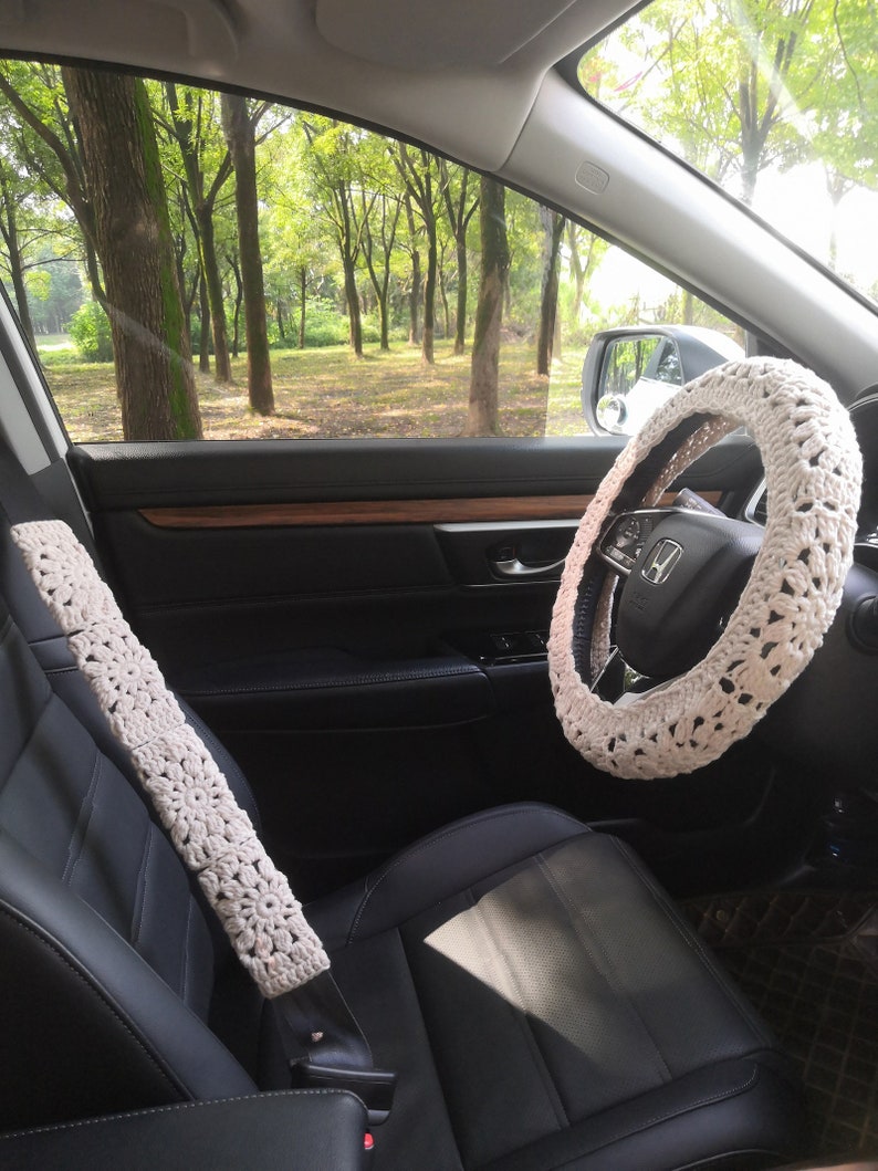 May include: A white crocheted steering wheel cover and a white crocheted seatbelt cover in a car interior. The car is parked in a wooded area.