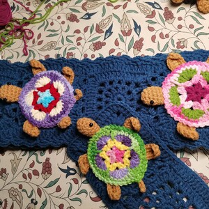 May include: Three crocheted turtle ornaments with colorful shell designs. The turtles are attached to a blue crocheted blanket.